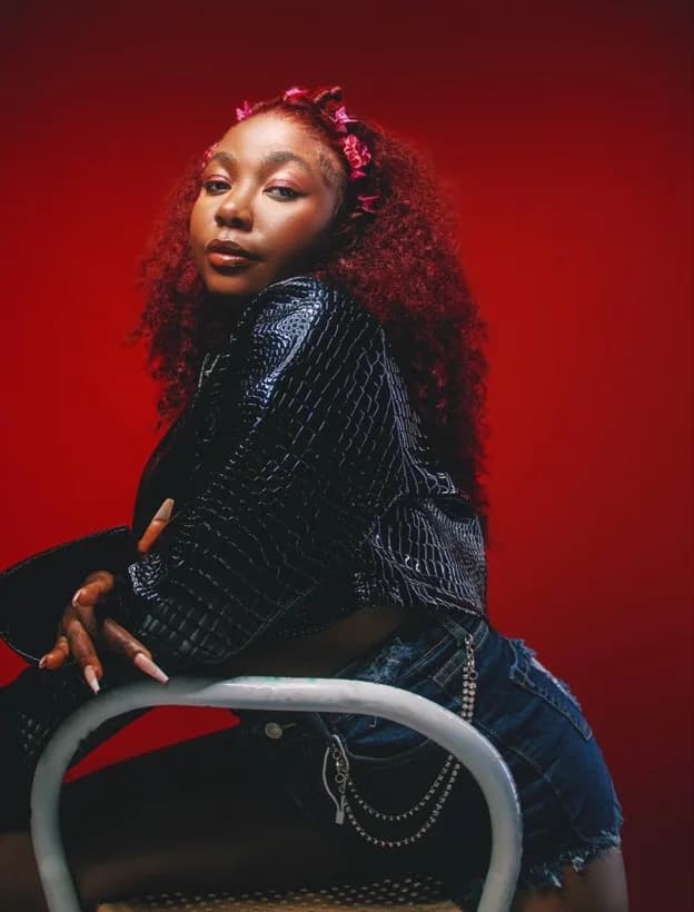 Big Jeanne seated on a chair in a black leather jacket against a red backdrop